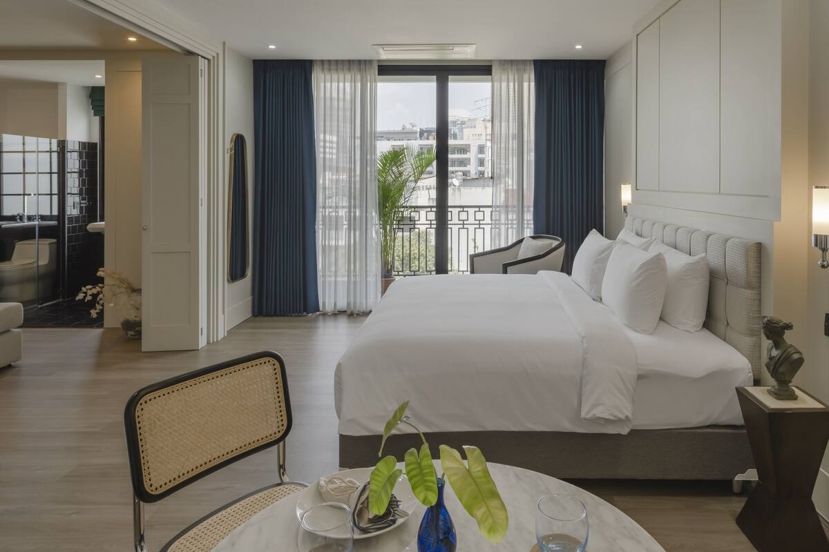 A hotel room with a large bed, a dining table with chairs, a balcony with a view of buildings, and a bathroom with a mirror and sink.