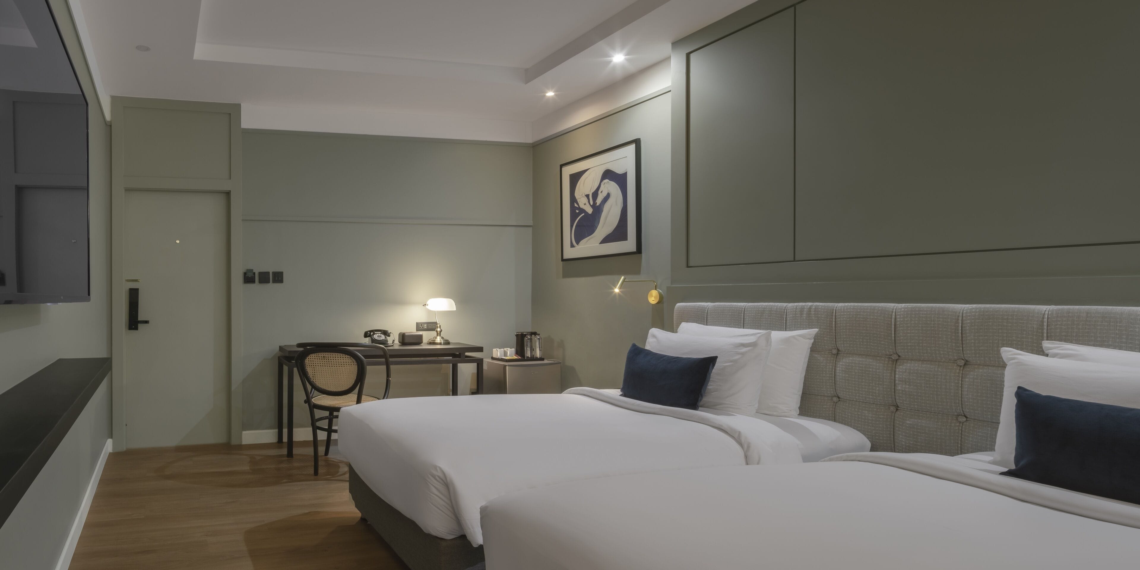 A hotel room with two beds, a desk, a chair, a lamp, and a framed artwork on the wall.