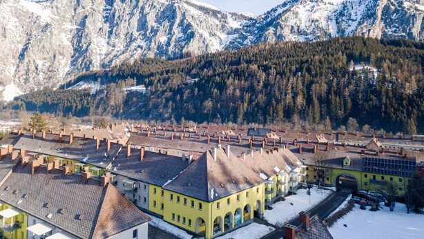 Un pueblo con arquitectura tradicional, un edificio central amarillo y montañas nevadas al fondo.