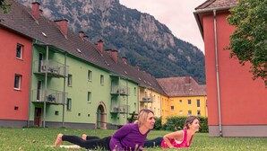 Dos personas realizando una postura de yoga en un césped frente a edificios coloridos, con una montaña al fondo.