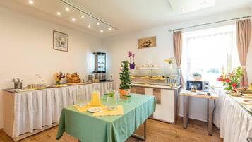 Un comedor con una mesa verde, mantel blanco y diversos alimentos. Hay dos refrigeradores, un microondas y una cafetera. El cuarto tiene pisos de madera y paredes blancas.