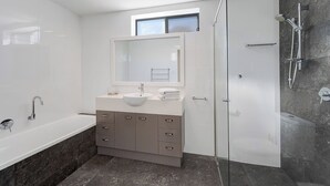 A modern bathroom with a bathtub, a sink with a mirror, a shower area, and a window. - Coochin Court | Walk to Dicky Beach (DICKY BEACH)
