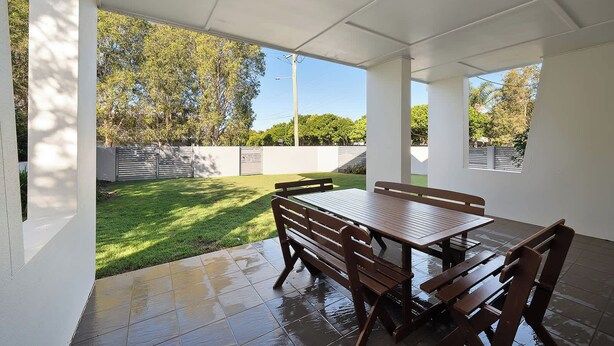 A covered patio with a table and chairs, a grassy lawn, and a white wall. - Coochin Court | Walk to Dicky Beach (DICKY BEACH)