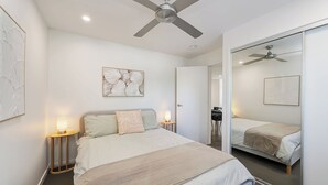 A modern bedroom with a bed, bedside lamps, a ceiling fan, and a large mirror. - Coochin Court | Walk to Dicky Beach (DICKY BEACH)