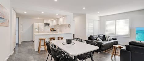 A modern apartment with a dining area, kitchen, living room, and a TV displaying a beach scene.