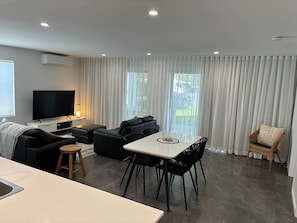 Un salon moderne doté d’un téléviseur à écran plat, d’un coin repas avec une table et des chaises, et d’une grande fenêtre agrémentée de rideaux.