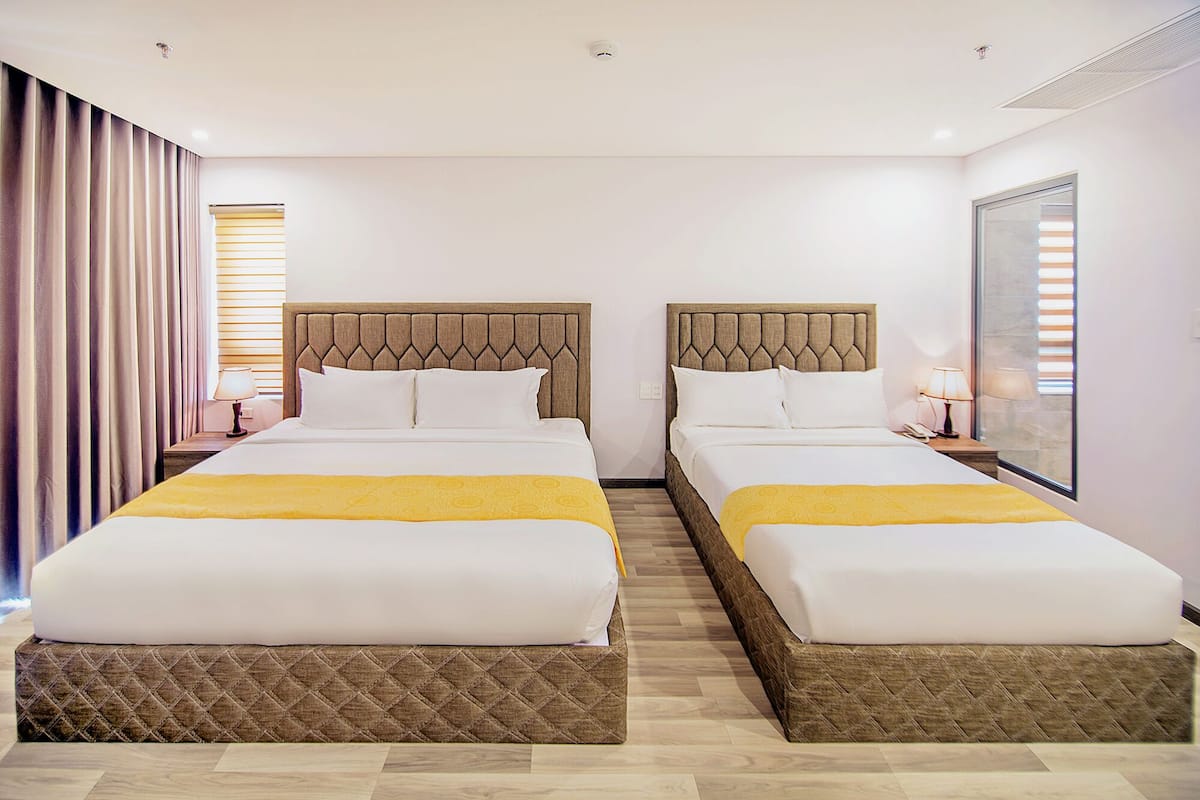 A hotel room with two beds, each with a wooden headboard, white bedding, and a yellow throw. There are bedside tables with lamps, a large window with blinds, and a curtain on the left.