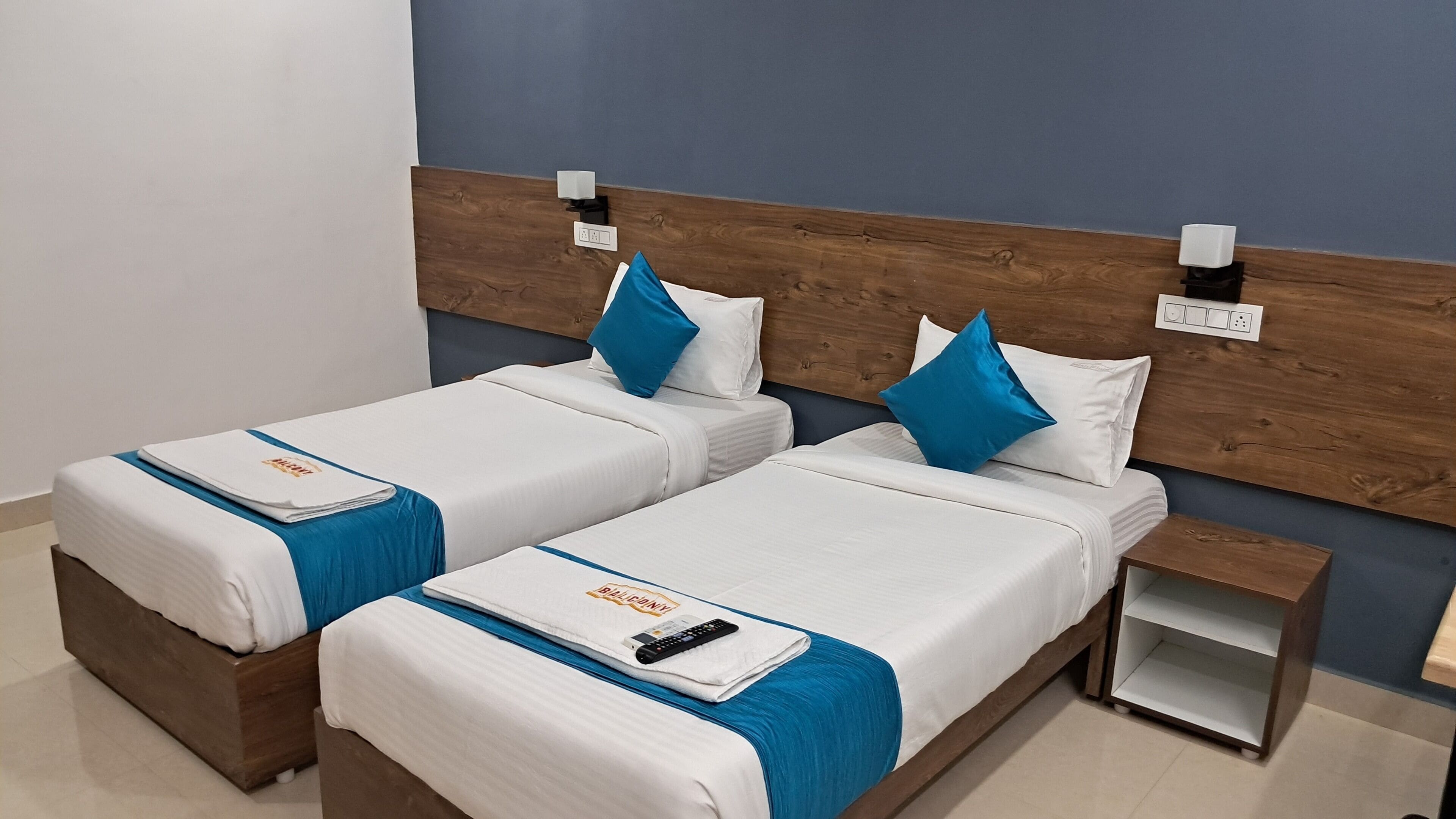 A hotel room with two beds, each with a blue and white bedspread, a remote control, and a small nightstand with a lamp.
