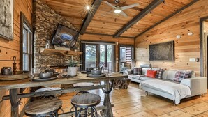 A rustic cabin interior with a stone fireplace, wooden dining table, bar stools, a sofa, and a ceiling fan. - Cherry Log Sky Bear | Cherry Log, GA (Blue Ridge)