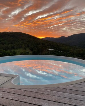 Un bain à remous sur une terrasse en bois, avec une vue sur le coucher de soleil et une colline boisée.