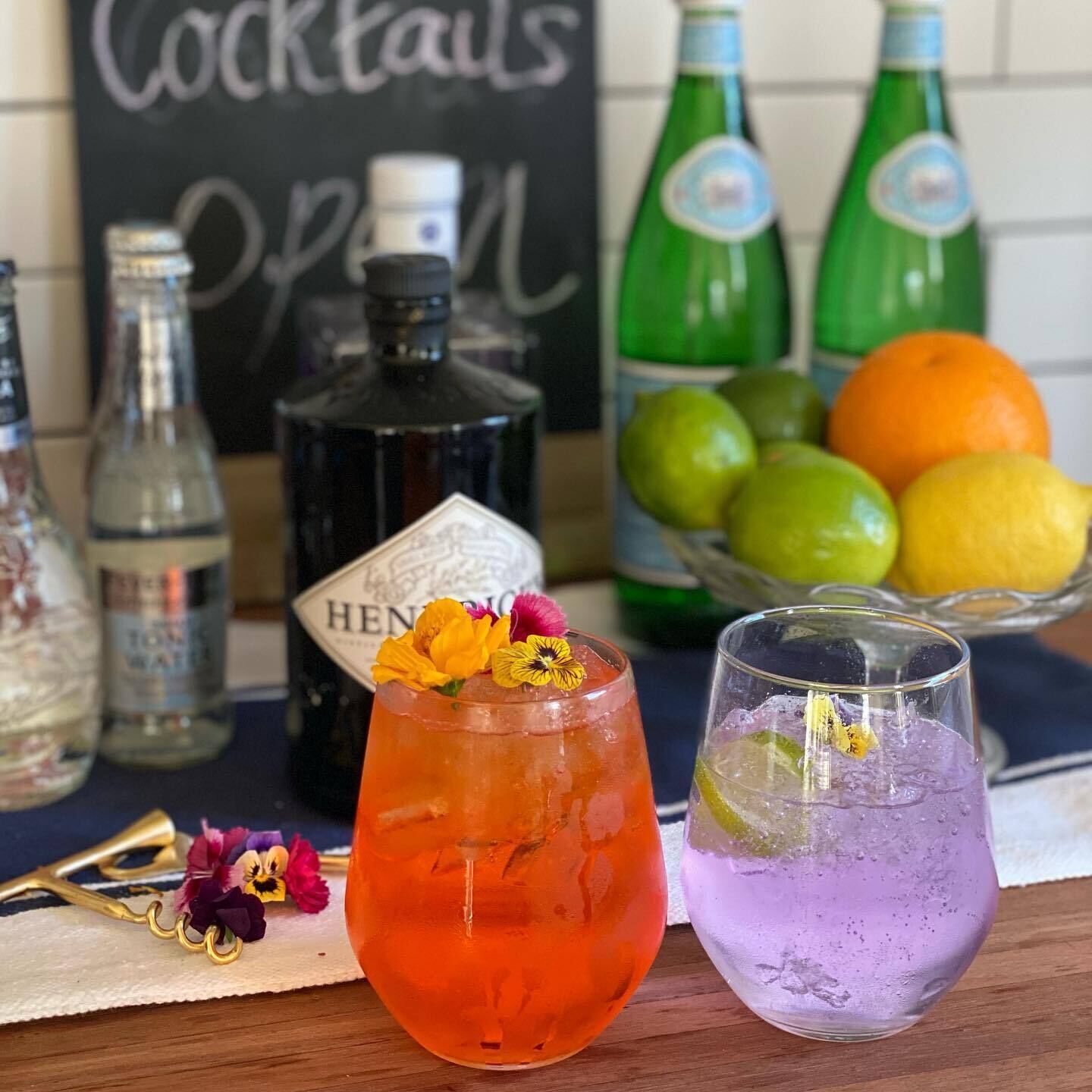 Two cocktails with flowers, a bottle of Hendrick's, and a bowl of fruits on a table.