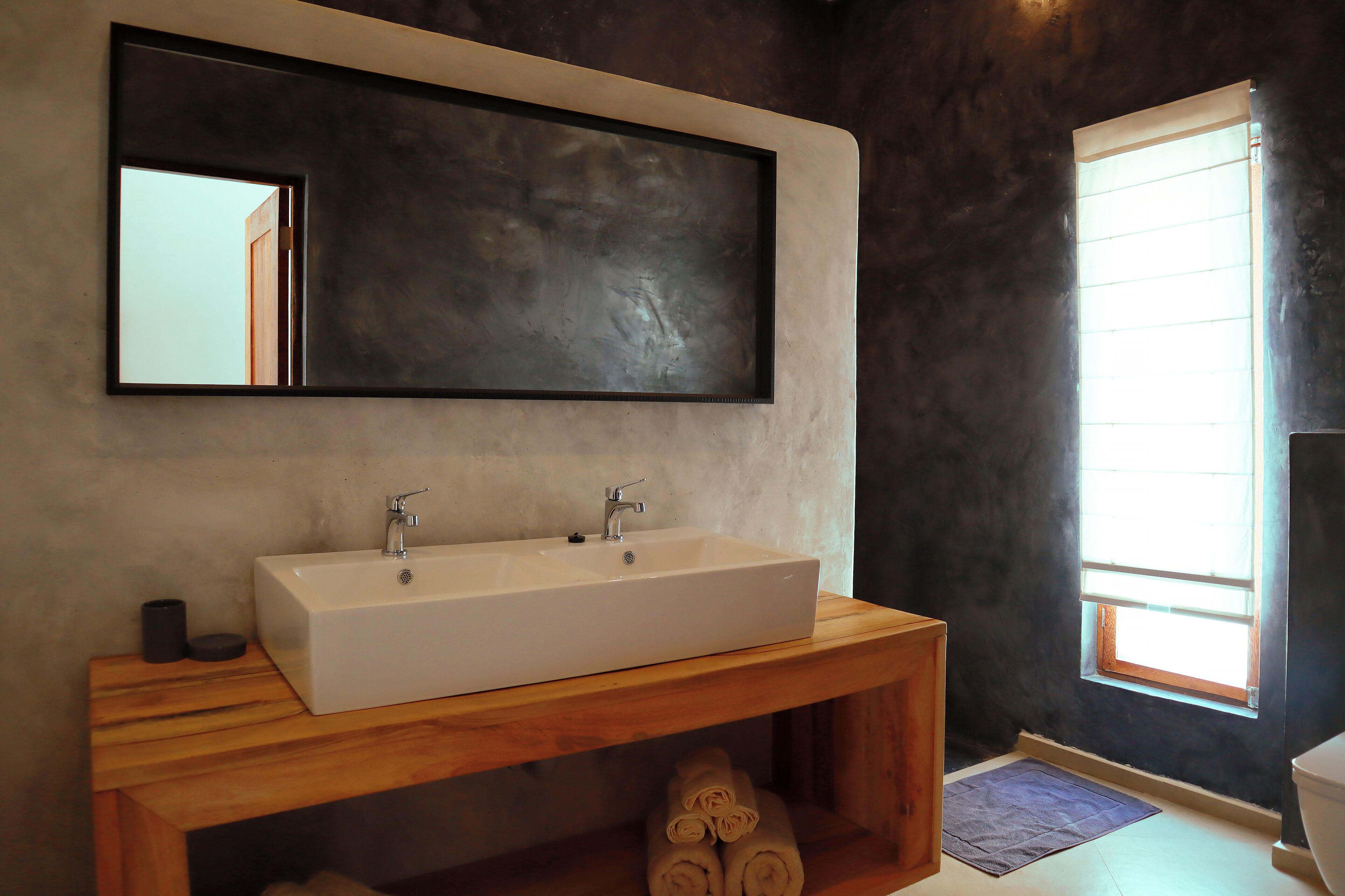 Un baño con un lavamanos grande y rectangular, un mueble de tocador de madera, un espejo y una ventana con persianas.