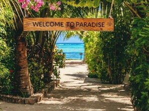 Uma placa de madeira com a inscrição 'WELCOME TO PARADISE', com um caminho que leva a uma praia.