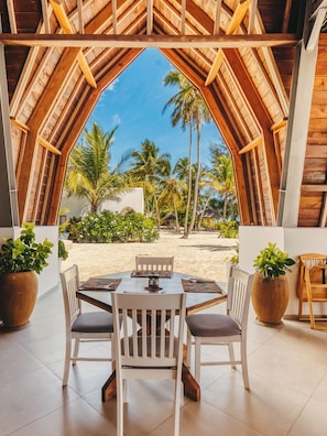 A dining area with a view of a beach and palm trees, featuring a wooden ceiling and white chairs. - Kijani Beach Villas (Bwejuu)