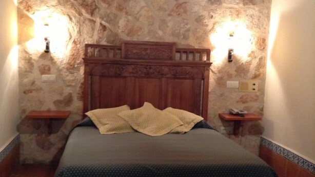 A bedroom with a stone wall, a wooden headboard, two bedside lamps, and a bed with a green bedspread. - Meson Don Quijote (Mota del Cuervo)