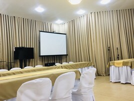 Sala de conferência com uma tela de projeção grande, cadeiras brancas e uma mesa com toalha.