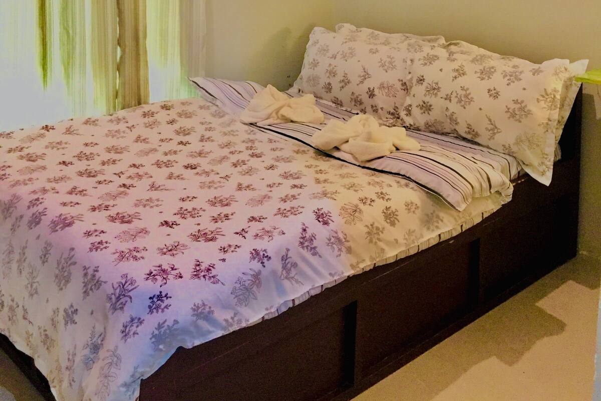 Un dormitorio con una cama, una ventana con cortinas, un equipo de aire acondicionado y dos cuadros colgados en la pared.