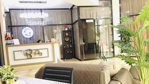 Eine Hotelhalle mit einer Rezeption, einer Sitzecke mit Tisch und Stühlen sowie einer Glastür zu einem weiteren Raum.
