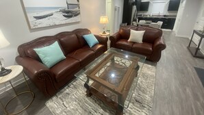 A living room with a brown leather sofa, a glass coffee table, and a painting of a beach scene. - BonVoyage-Sleep&Sail-OvernightLayovers-Sleeps 8 Near Airport-Cruise Ports (Long Beach)