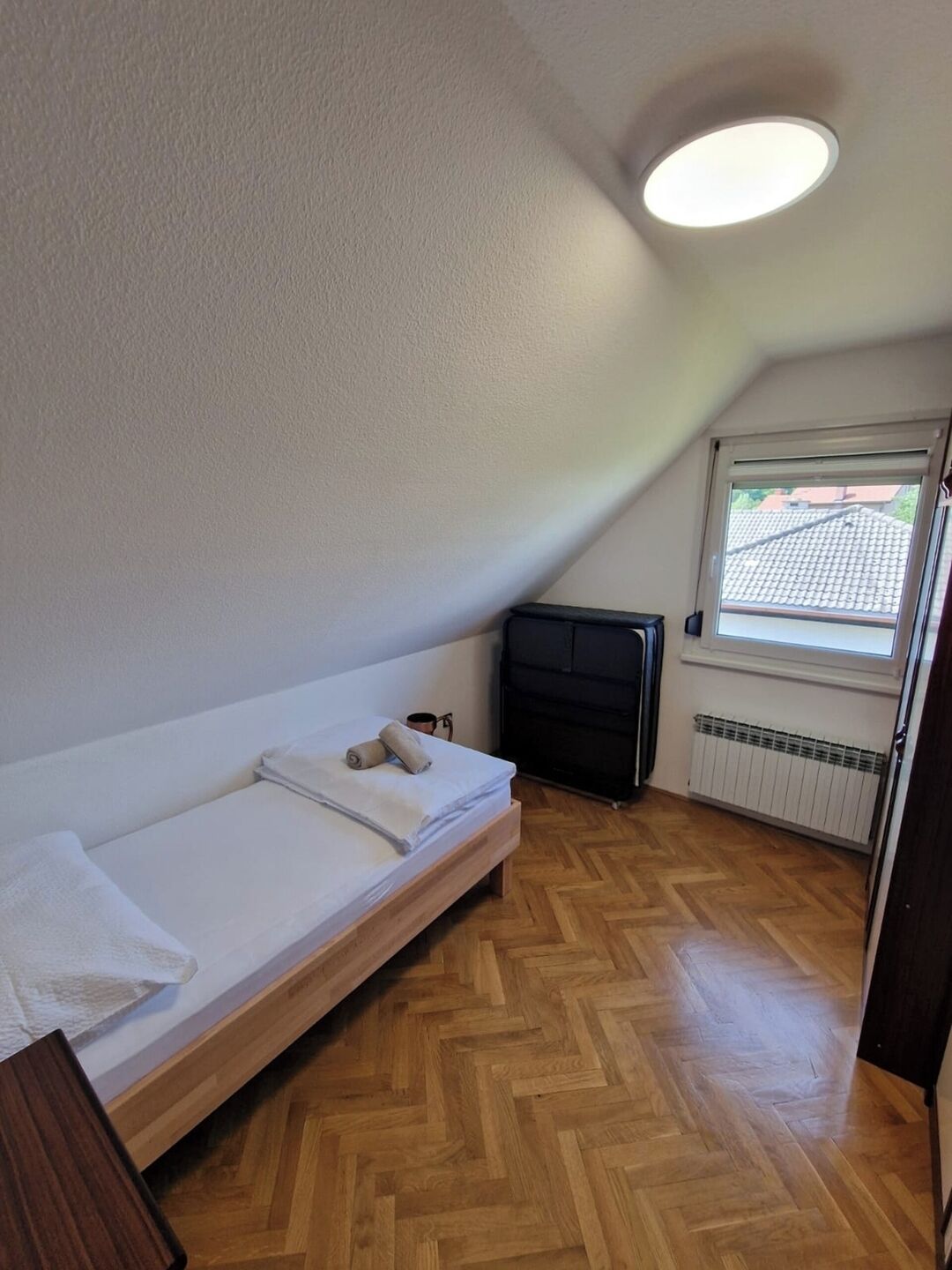 Un petit appartement d’une seule pièce, avec un parquet en bois disposé en chevrons, un lit, un radiateur et une fenêtre donnant sur l’extérieur.