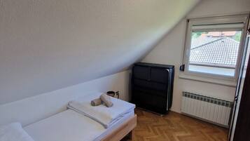 Un petit appartement d’une seule pièce, avec un parquet en bois disposé en chevrons, un lit, un radiateur et une fenêtre donnant sur l’extérieur.