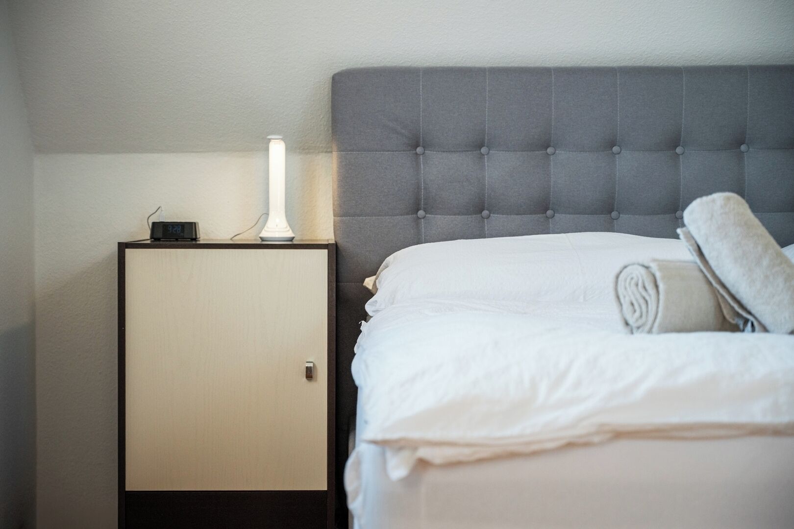 A neatly made bed with a grey tufted headboard, a bedside table with a lamp, and a digital clock.