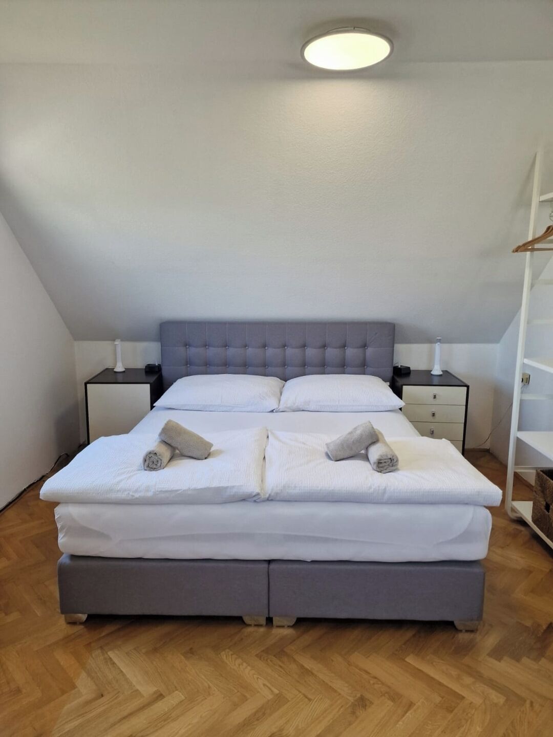 A bedroom with a bed, two bedside tables, a nightstand, a ceiling light, and a wooden floor.