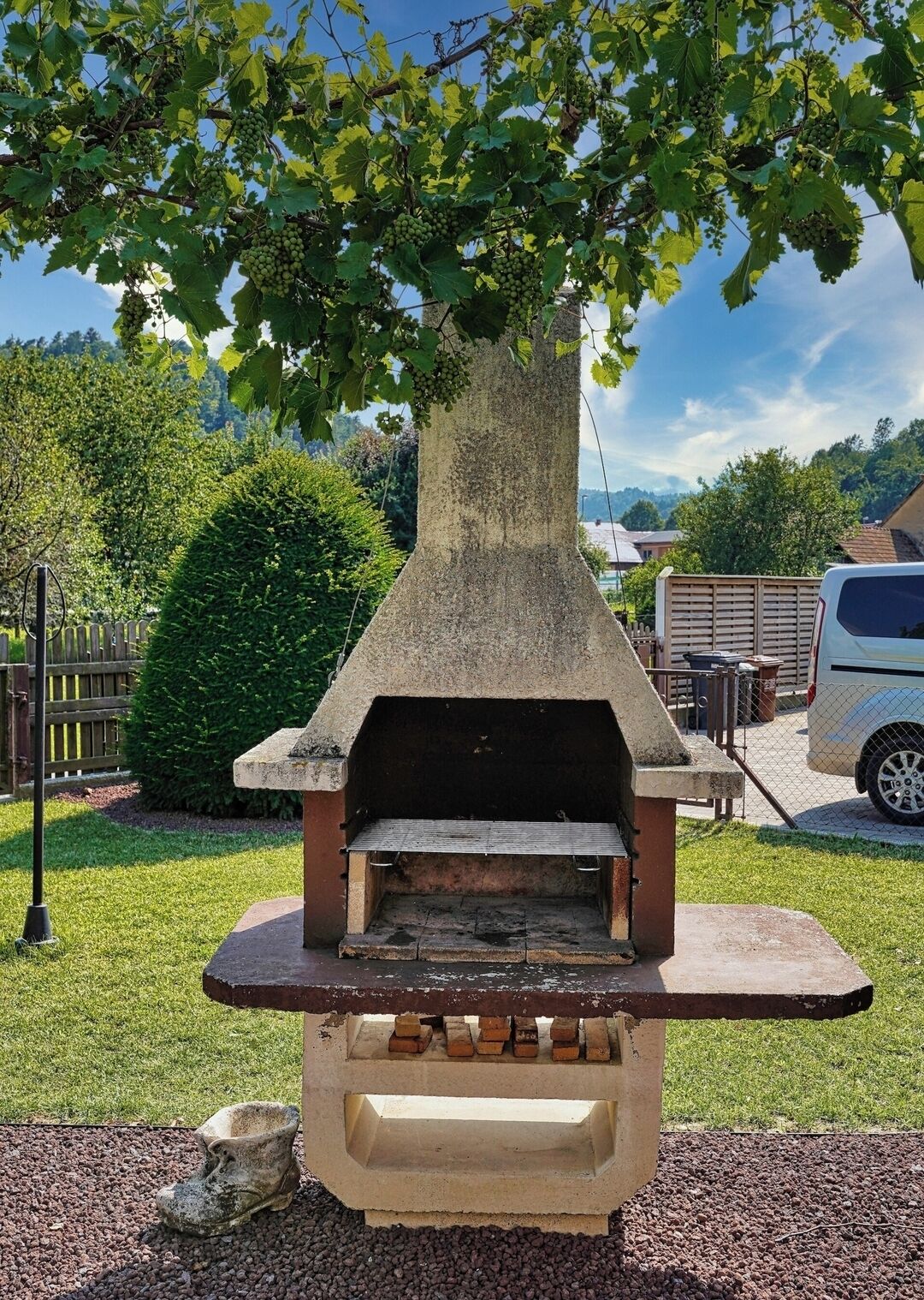 Una barbacoa exterior de formigó amb xemeneia, envoltada d'una zona verda i un arbre.