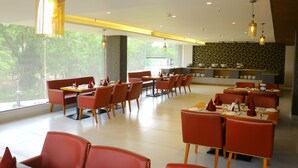 A modern restaurant with red chairs and tables, large windows, and a kitchen area. - Tulip Arena Hotel Club Event By SGL (Raipur)