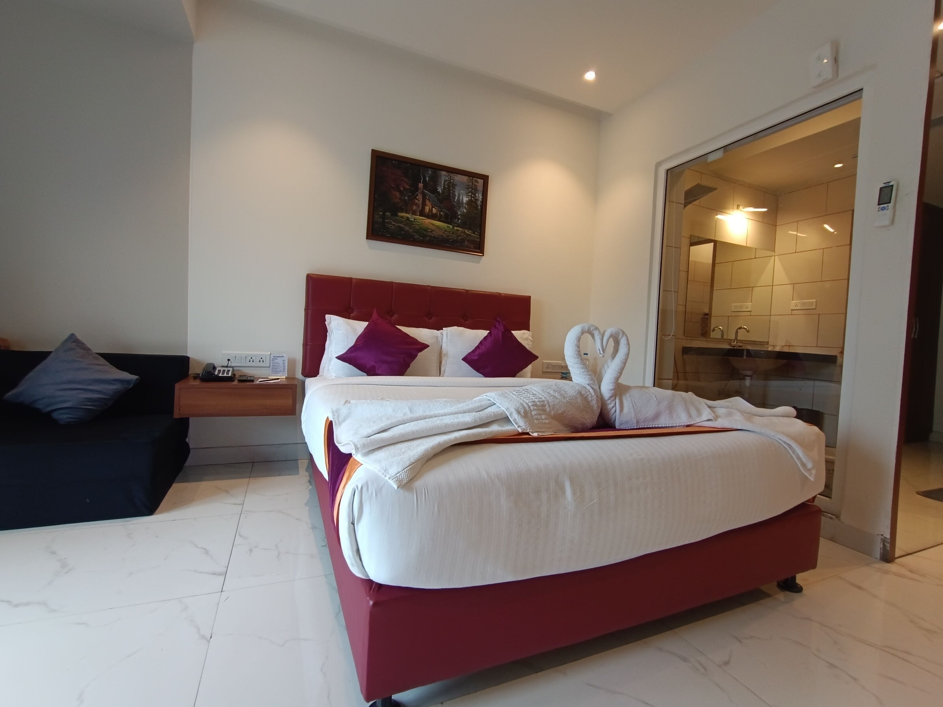 Una habitación de hotel con una cama grande, una mesita de noche, un cuadro en la pared y un baño visible a través de una puerta abierta.