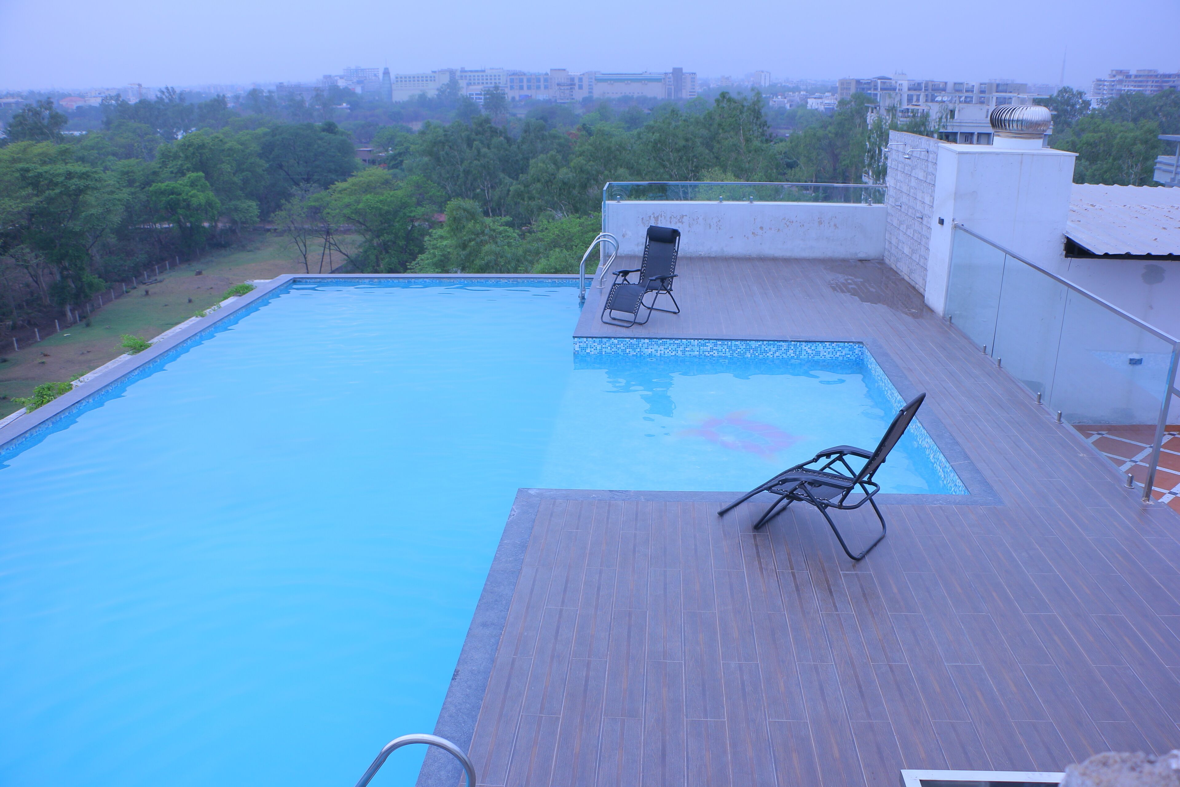A rooftop swimming pool with a wooden deck, a lounge chair, and a view of the cityscape.