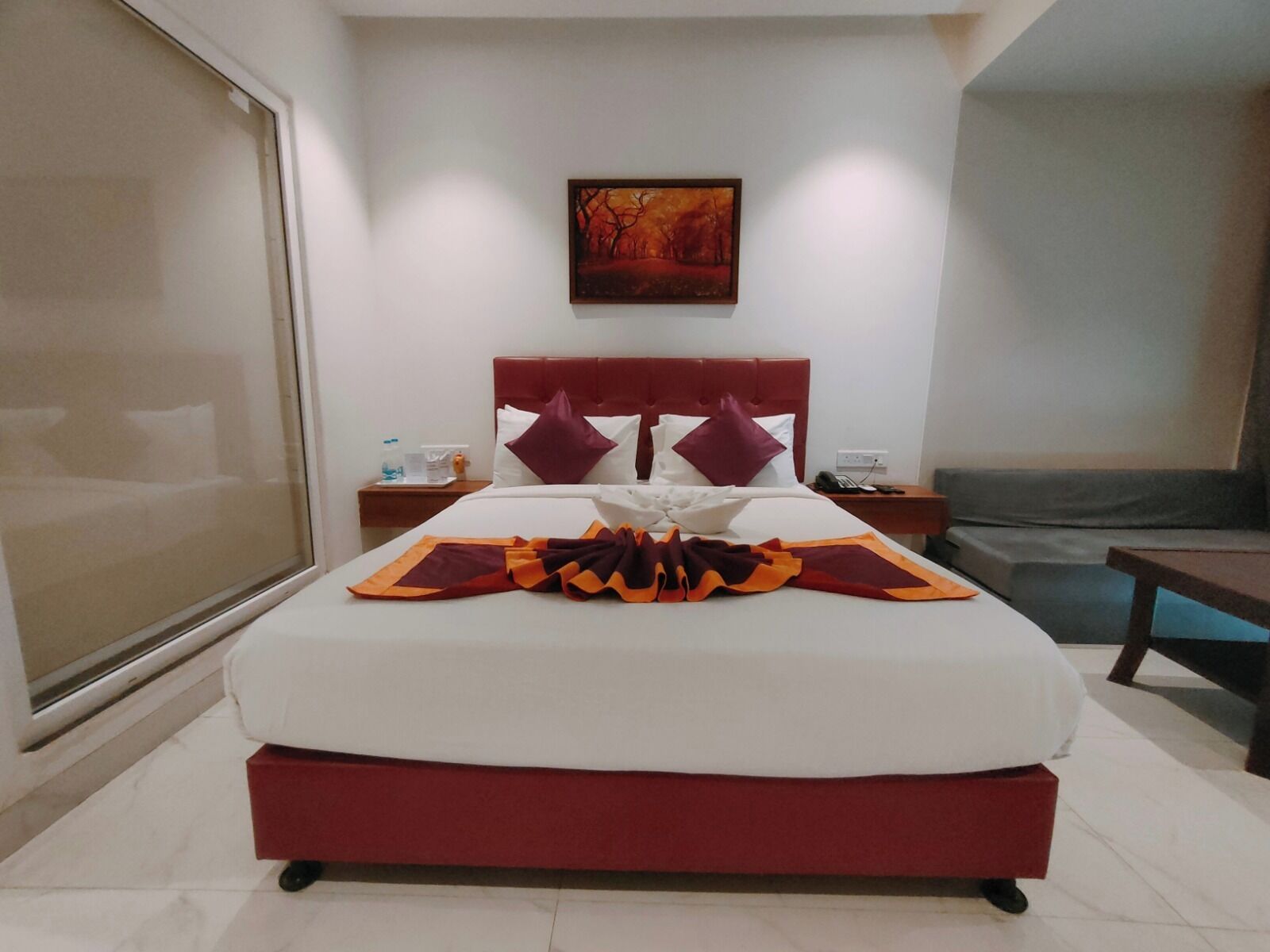 Une chambre d’hôtel avec un grand lit, une tête de lit, une table de chevet, une commode, une chaise, un canapé, un tableau au mur et une fenêtre.