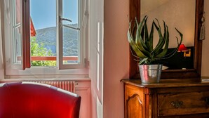 Una stanza con una sedia rossa, un comò in legno, una pianta in vaso e una finestra che si affaccia su vegetazione e colline.