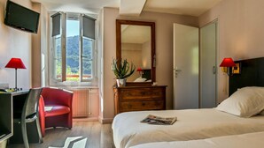 Una camera d'albergo con un letto, una scrivania con una sedia, una TV, un comò, uno specchio e una finestra vista alberi.