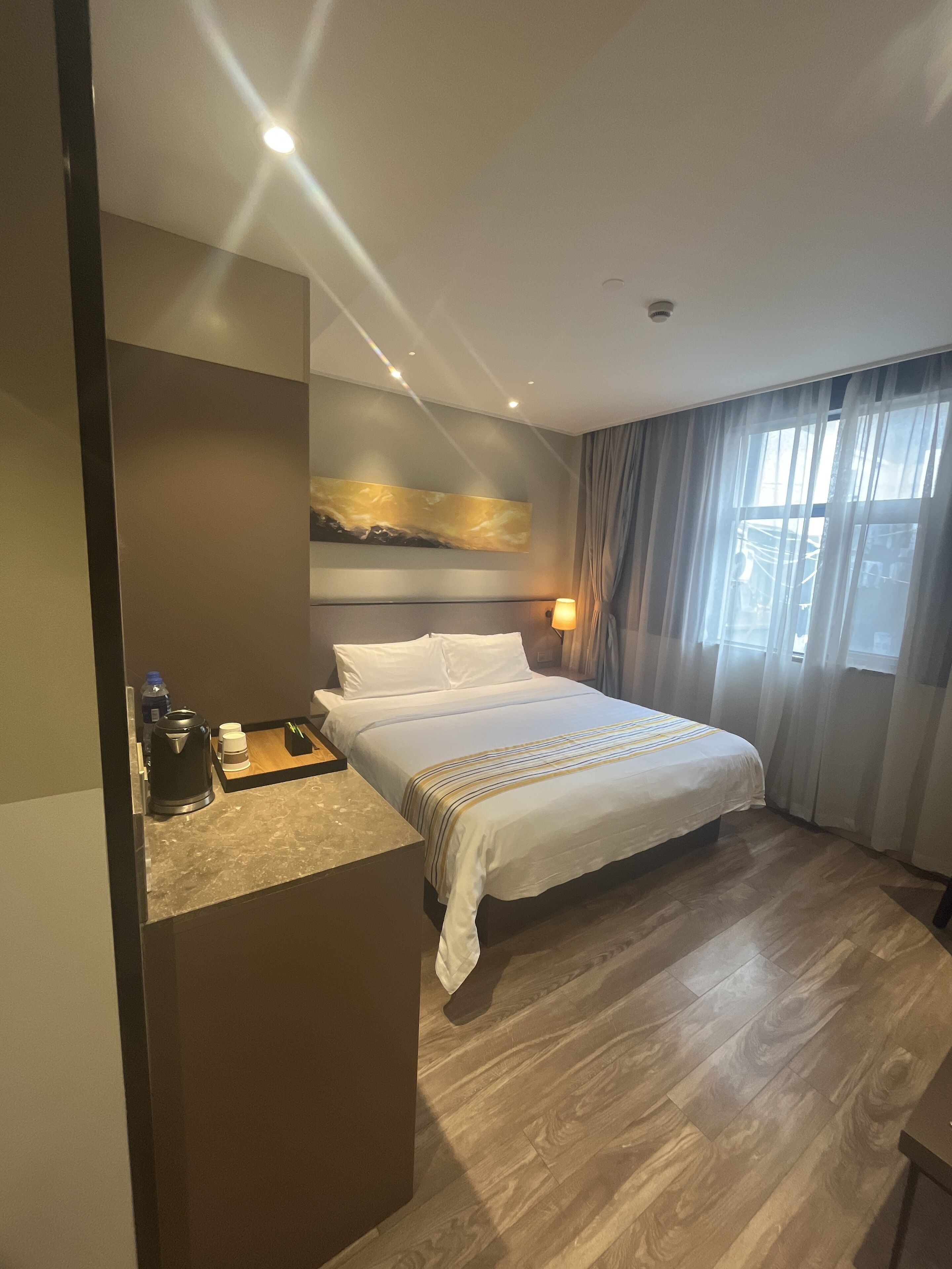 A hotel room with a bed, a bedside table, a lamp, a painting on the wall, and a window with curtains.
