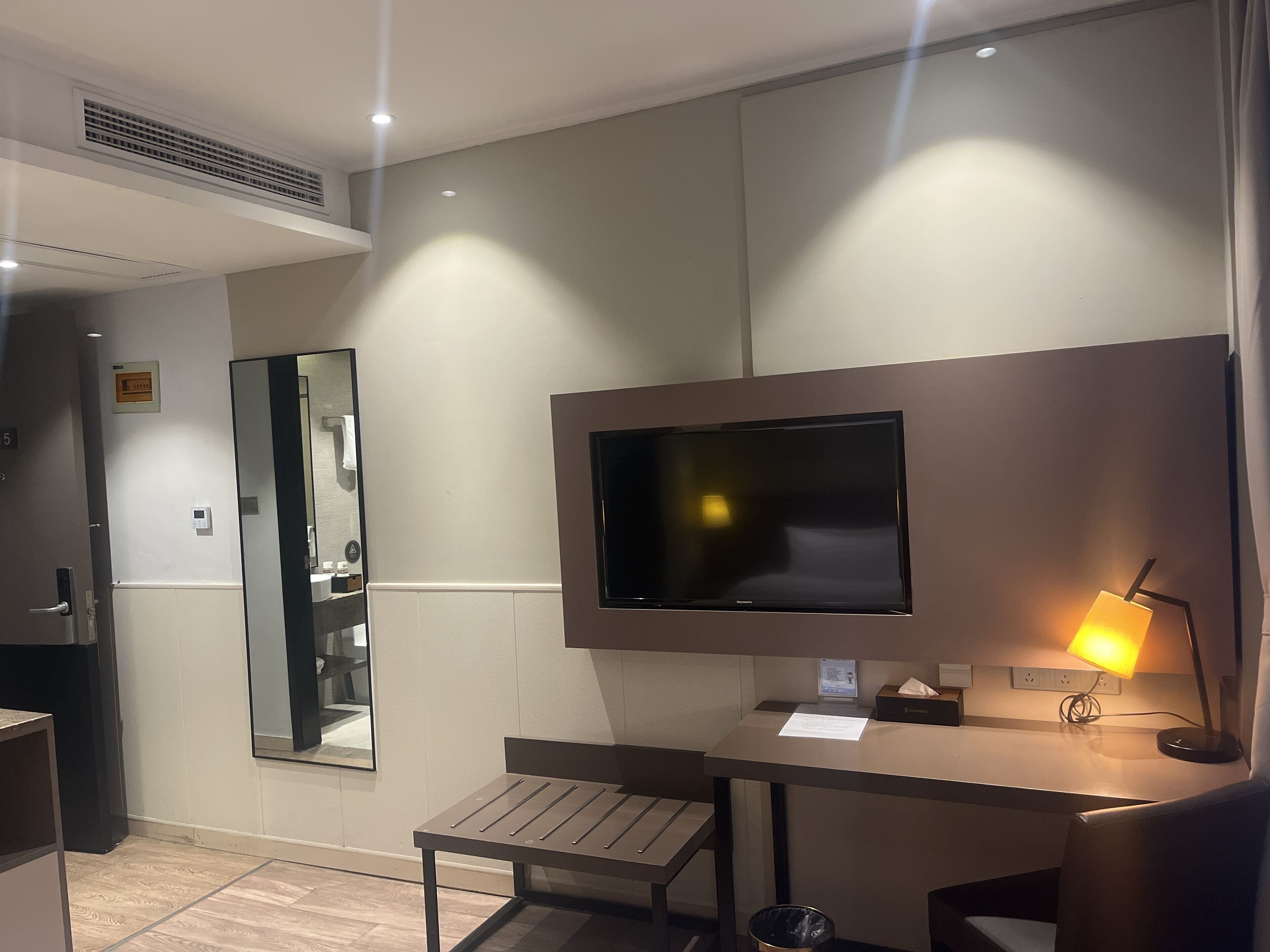 A modern hotel room with a flat-screen TV mounted on the wall, a small desk with a lamp, a bench, and a trash can.