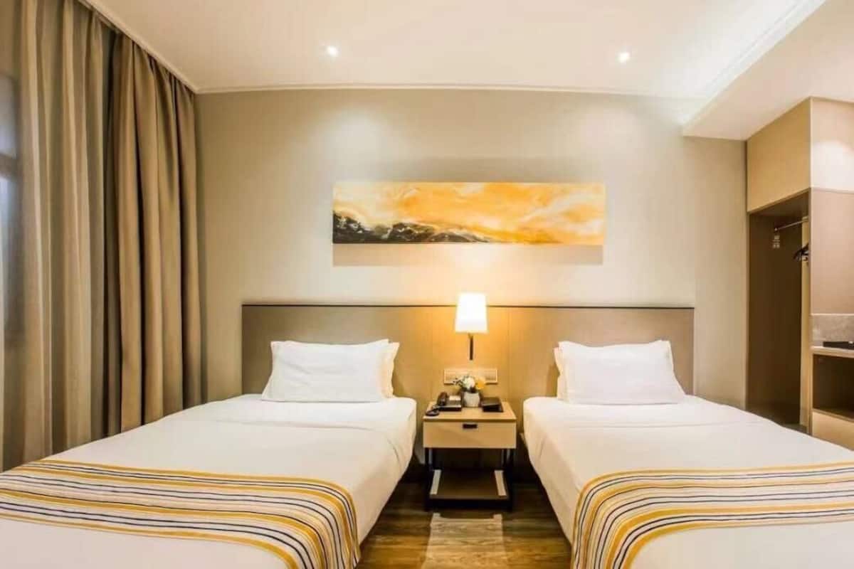 A hotel room with two beds, a nightstand with a lamp, a painting on the wall, and a wardrobe.