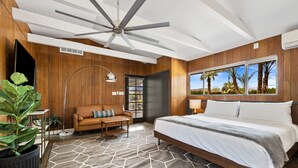 A modern bedroom with a large bed, a ceiling fan, a wooden wall panel, a window with a view of palm trees, and a geometric rug. - The Long Ranch - Sprawling 1 acre Horse Park Hideaway (Thermal)