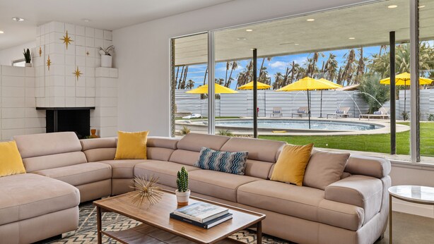 A modern living room with a beige sectional sofa, a wooden coffee table, and a fireplace. Large windows offer a view of a pool area with yellow umbrellas. - The Long Ranch - Sprawling 1 acre Horse Park Hideaway (Thermal)