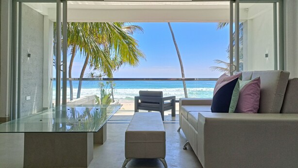 Una sala de estar moderna con una mesa de vidrio, un sofá con cojines y vista al océano y palmeras.