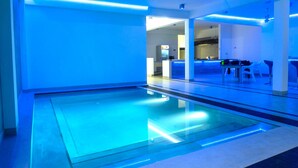 Piscina interior moderna con iluminación azul, zona de recepción y área de descanso con mesas y sillas.