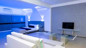 Una habitación de hotel moderna con una cama, sofás blancos, una mesa de centro de vidrio y un televisor de pantalla plana montado en la pared.