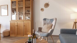 Ein Wohnzimmer mit einem Holzschrank, einem blauen Sessel, einem runden Holztisch mit einer Flasche und Gläsern sowie einer kleinen Vase mit Blumen.