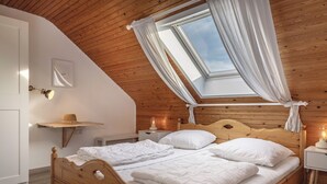 Ein Schlafzimmer mit Holzbalkendecke, einem Oberlicht mit weißen Vorhängen, einem hölzernen Bett mit weißer Bettwäsche, einem Nachttisch mit Lampe und einem Wandregal mit einem Hut.