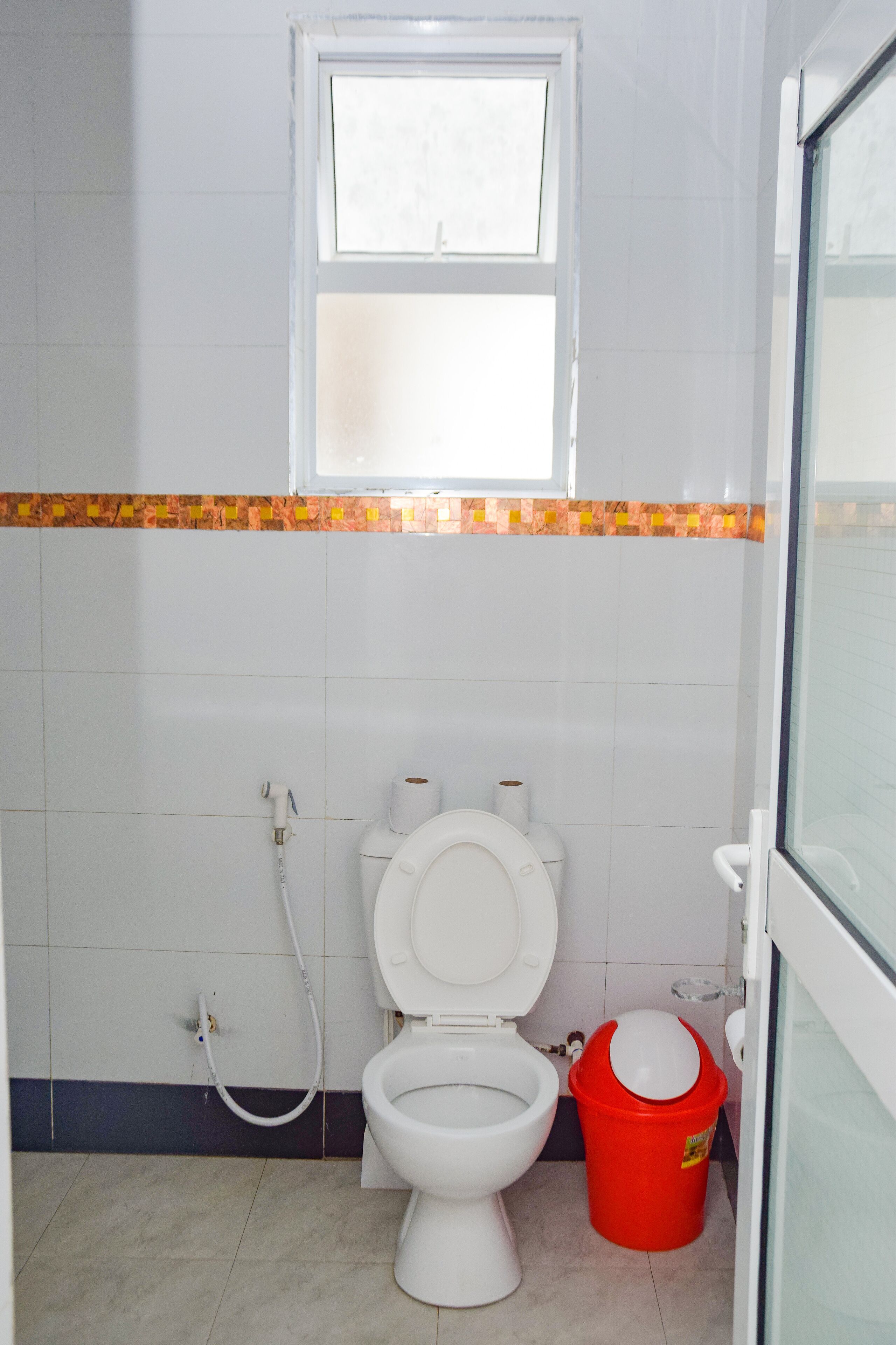 Un baño con inodoro, ventana y un balde rojo.
