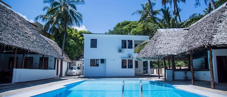 A swimming pool with a thatched-roof structure on one side and a white building in the center.