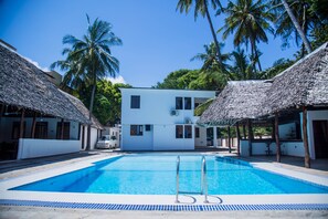 A swimming pool with a thatched-roof structure on one side and a white building in the center.