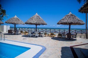 A poolside area with a clear view of the ocean, thatched umbrellas, and lounge chairs.