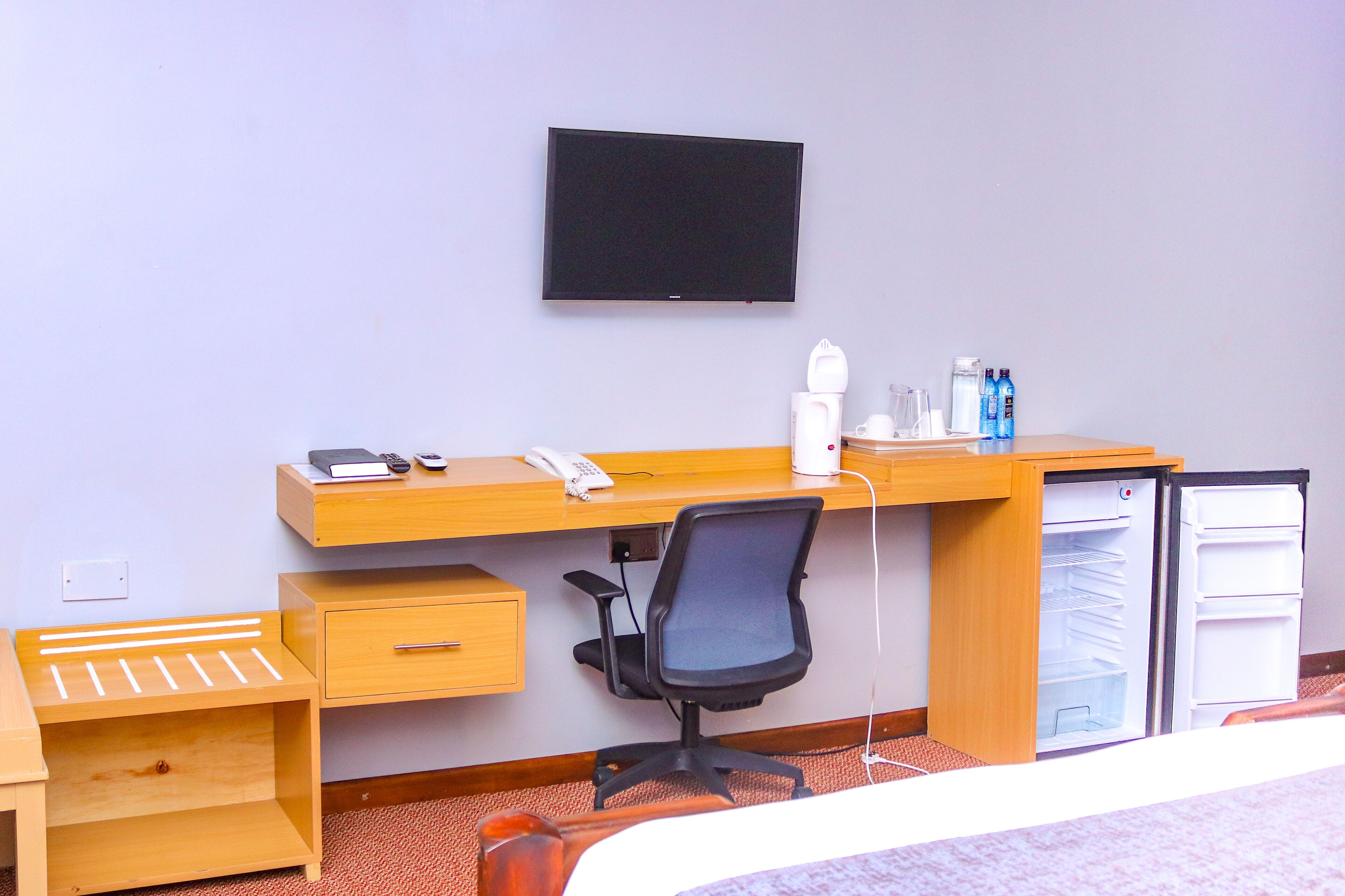 A hotel room with a wooden desk, a black office chair, a flat-screen TV mounted on the wall, a mini-fridge, and a small wooden side table with a lamp.