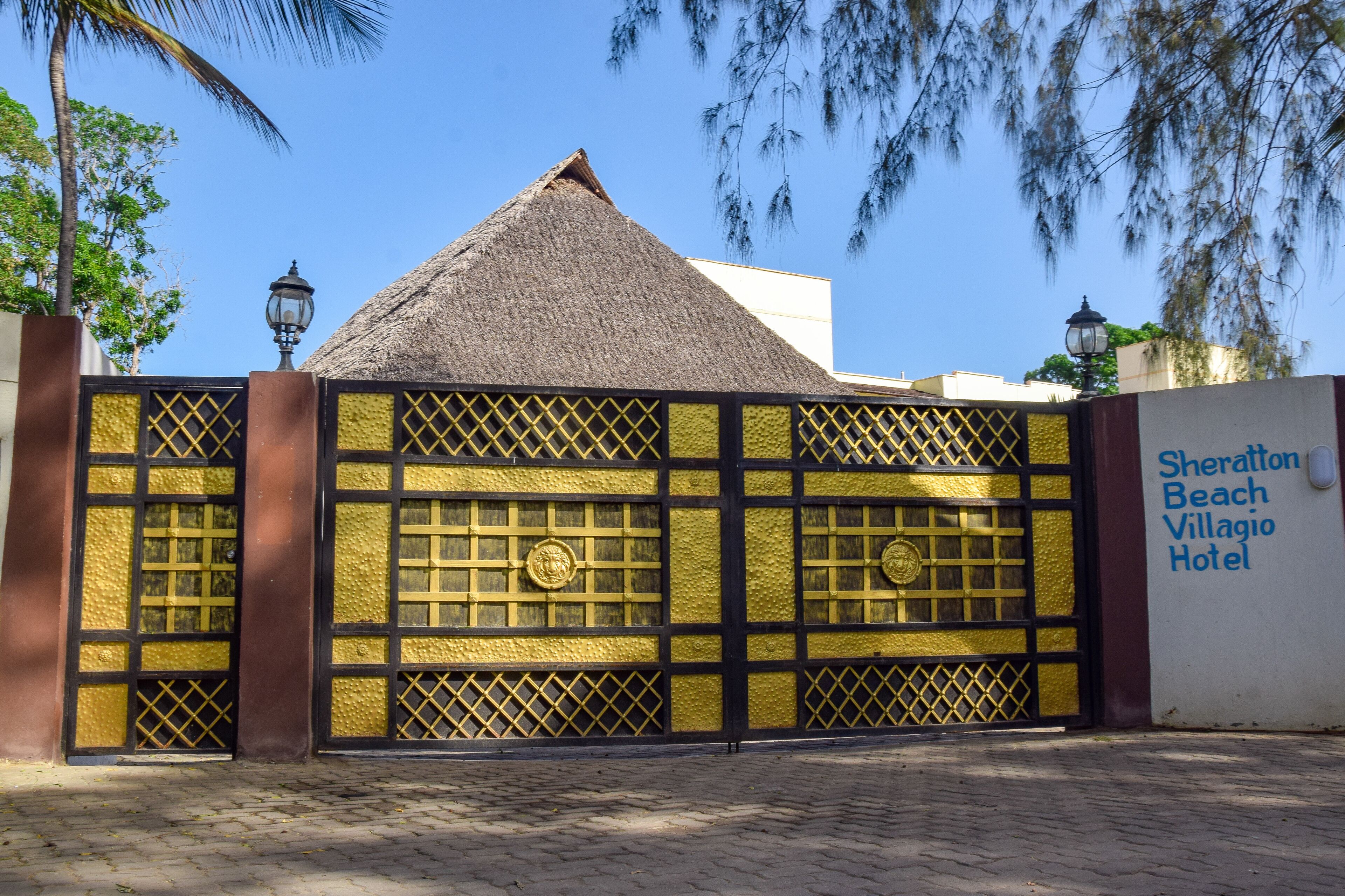 Een poort met een rieten dak, een decoratieve poort met gouden patronen, en een bord met de tekst Sheraton Beach Villagio Hotel.